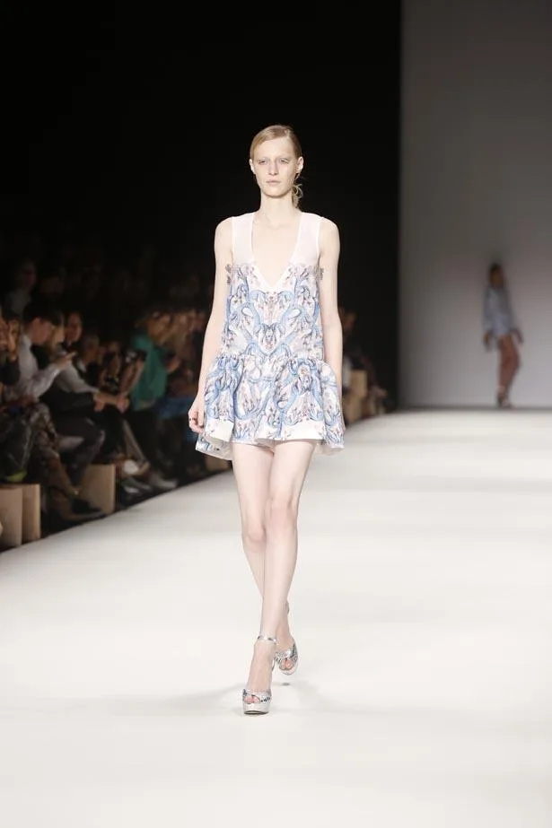 Model on runway wearing blue floral dress, Alice McCall SS13-14 collection. Audience visible in background.