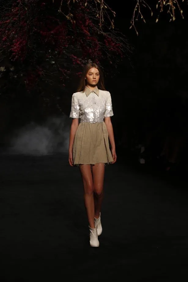 Model walking on a dimly lit runway, wearing a white blouse and tan skirt, with branches overhead.