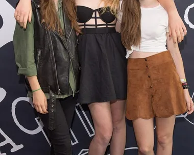 Three women, members of Haim, pose together in front of a backdrop, all wearing sunglasses and casual outfits.