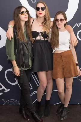 Three women, members of Haim, pose together in front of a backdrop, all wearing sunglasses and casual outfits.