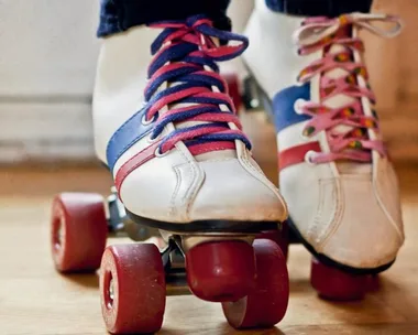 Close-up of two pairs of feet wearing white and blue roller skates with colorful laces, standing on a wooden floor.