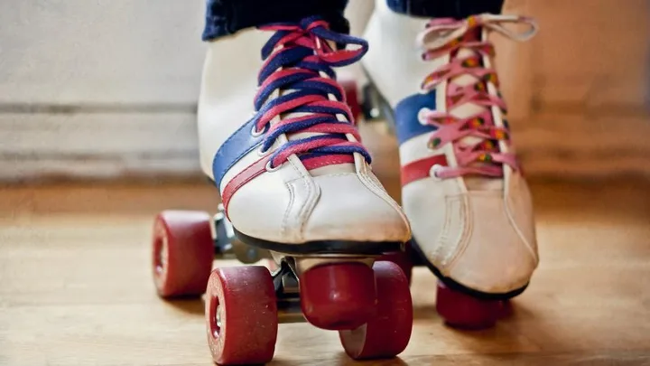 Close-up of two pairs of feet wearing white and blue roller skates with colorful laces, standing on a wooden floor.