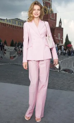 A woman in a light pink suit stands outdoors near a red brick building with a clock tower, holding a white bag.