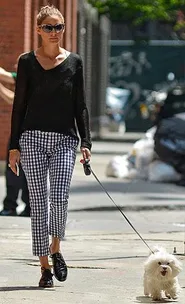 A woman walks a small white dog on a city street, wearing a black sweater and gingham pants.