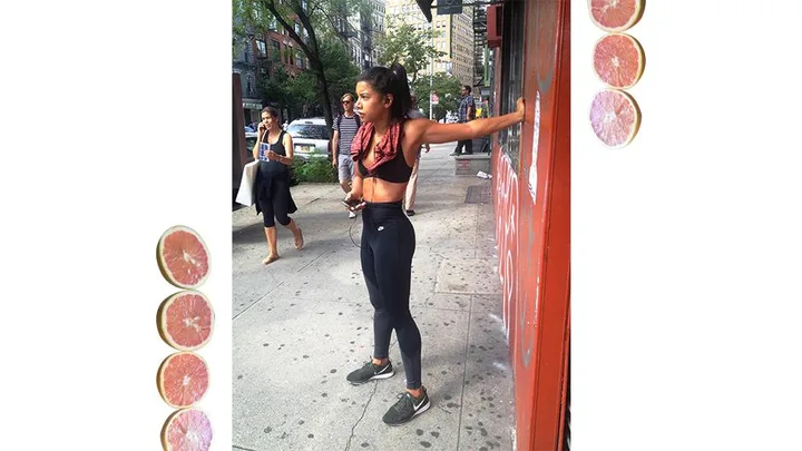 Woman in black athletic attire stretching on a city street, with a red towel around her neck and people walking in the background.