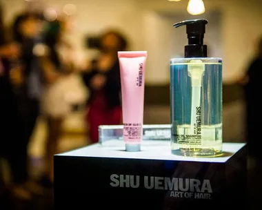 Shu Uemura hair products displayed on a stand with blurry people in the background.