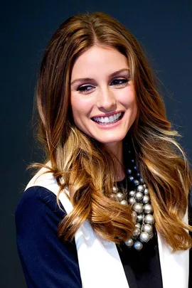 Woman with long wavy brown hair smiling, wearing a black and white outfit and a chunky silver necklace.