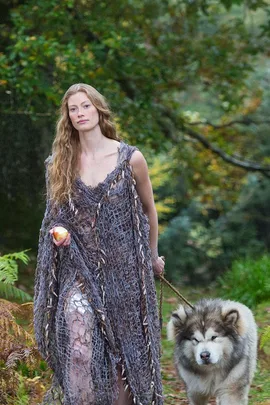 A woman in a draped mesh dress holds an apple and walks a large, fluffy dog in a forest setting.