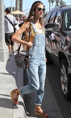 Woman in denim overalls and sunglasses walks on a sunny street, carrying a crossbody bag and wearing wedge sandals.