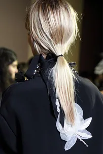 A person with blonde hair tied in a simple ponytail, wearing a dark top with white floral embellishments on the back.