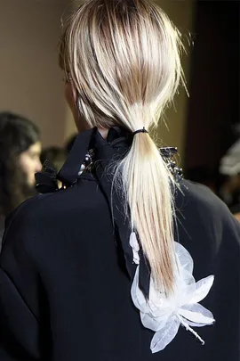 A person with blonde hair tied in a simple ponytail, wearing a dark top with white floral embellishments on the back.