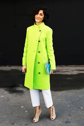 A person in a bright green coat, white pants, and beige heels holding a book, smiles against a dark background.