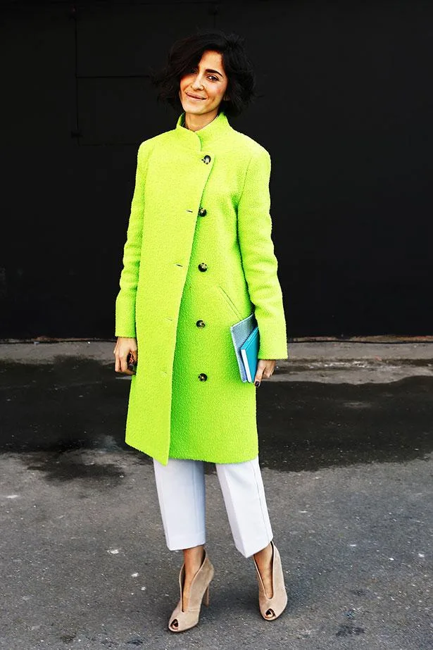 A person in a bright green coat, white pants, and beige heels holding a book, smiles against a dark background.