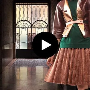 Fashionable person in a pleated skirt and jacket stands inside a grand corridor. Play button overlay indicates a video.