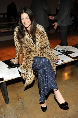 Woman in leopard print coat and polka dot pants sitting on a bench, smiling at a fashion event.