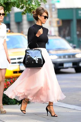 Woman in black top and pink skirt walks on city street carrying a large black handbag.