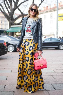 Woman in a black leather jacket, graphic tee, and yellow pattern skirt holds a bright pink handbag at Milan street style event.