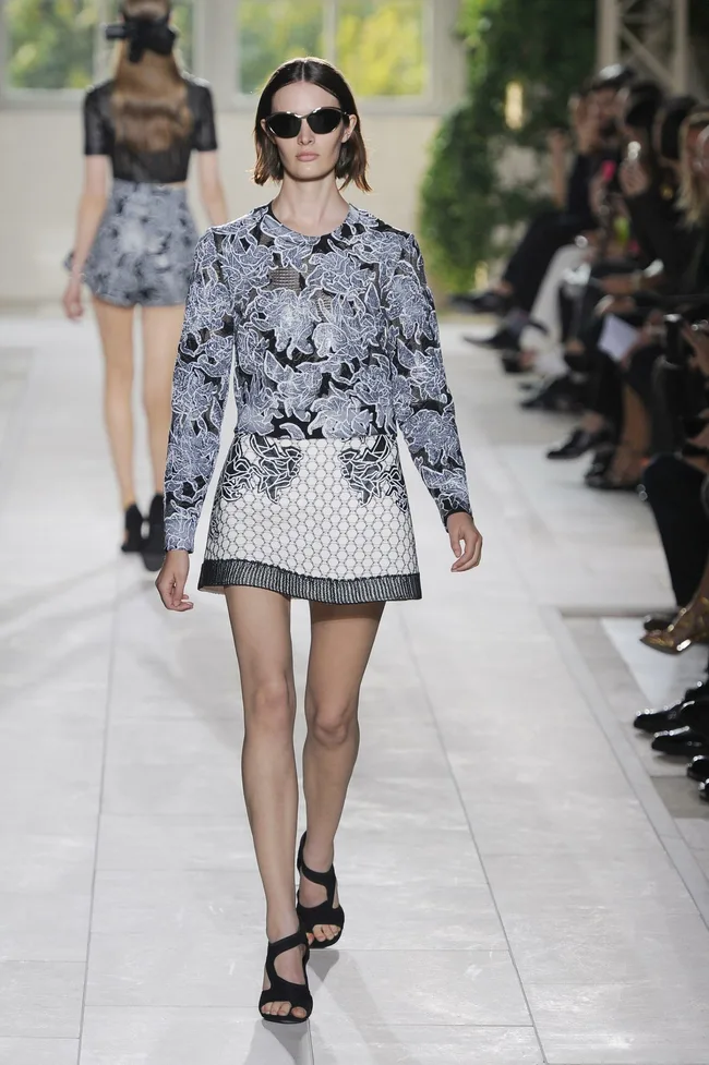Model walks runway in black and white floral outfit with sunglasses, surrounded by seated audience.