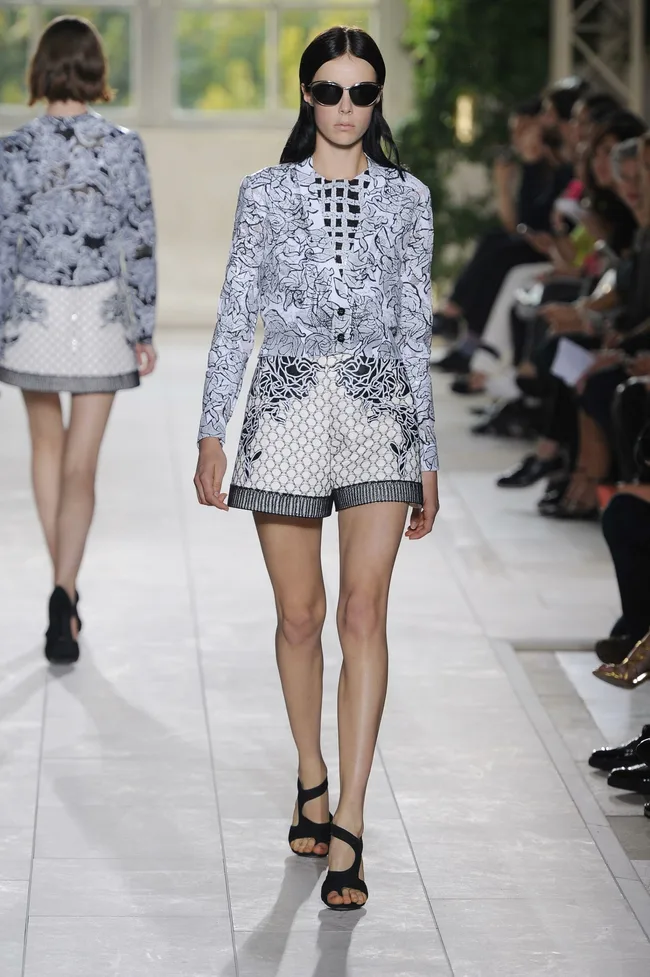 Model in patterned outfit and sunglasses walking the runway, with audience watching in the background.