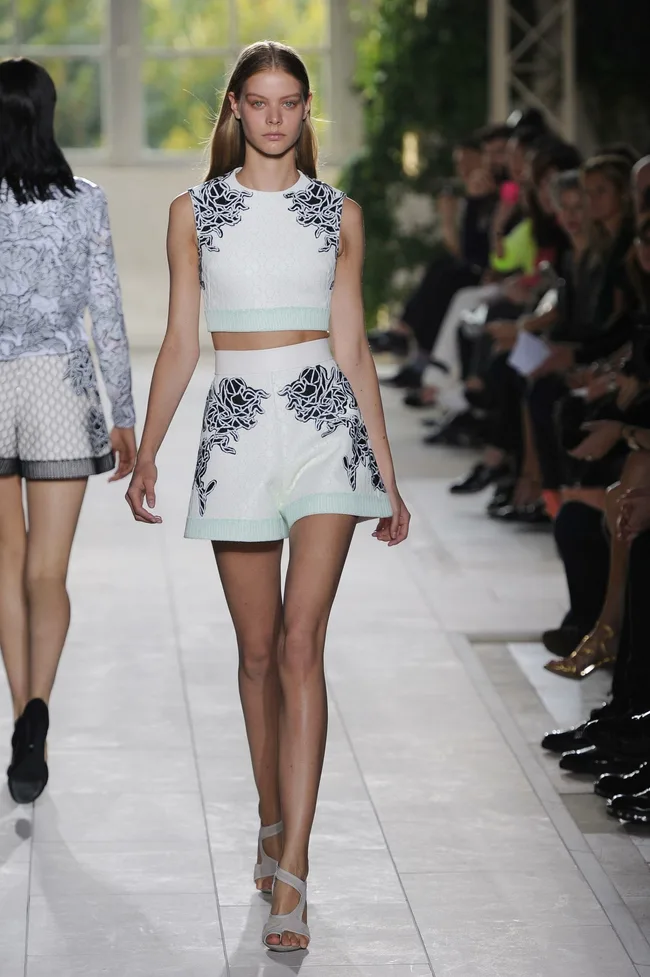 Model walking runway in a white crop top and shorts with black floral patterns at a fashion show.