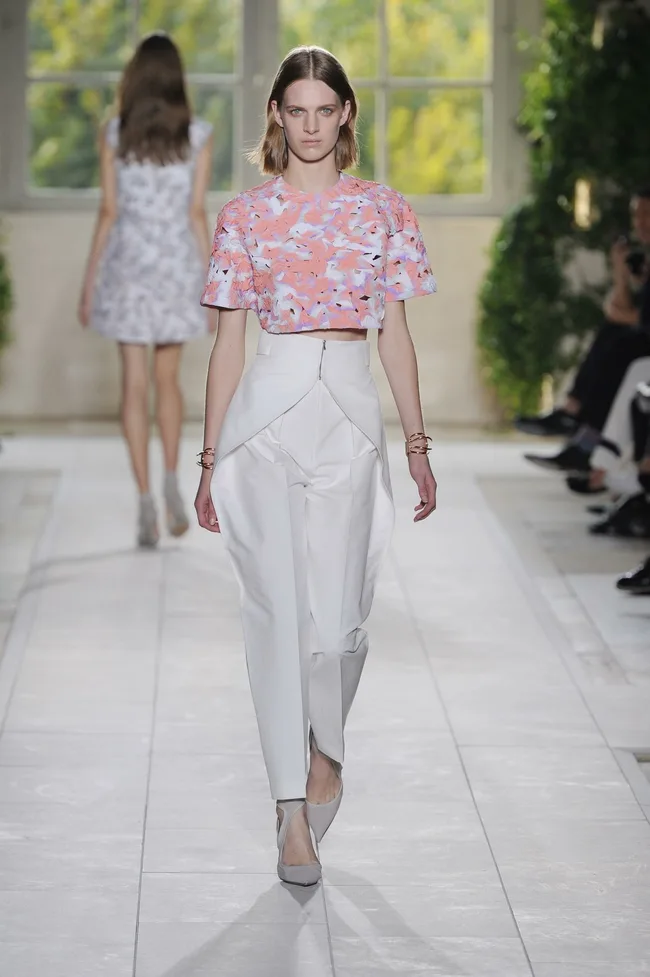 Model in floral crop top and white pants walks runway with leafy background.