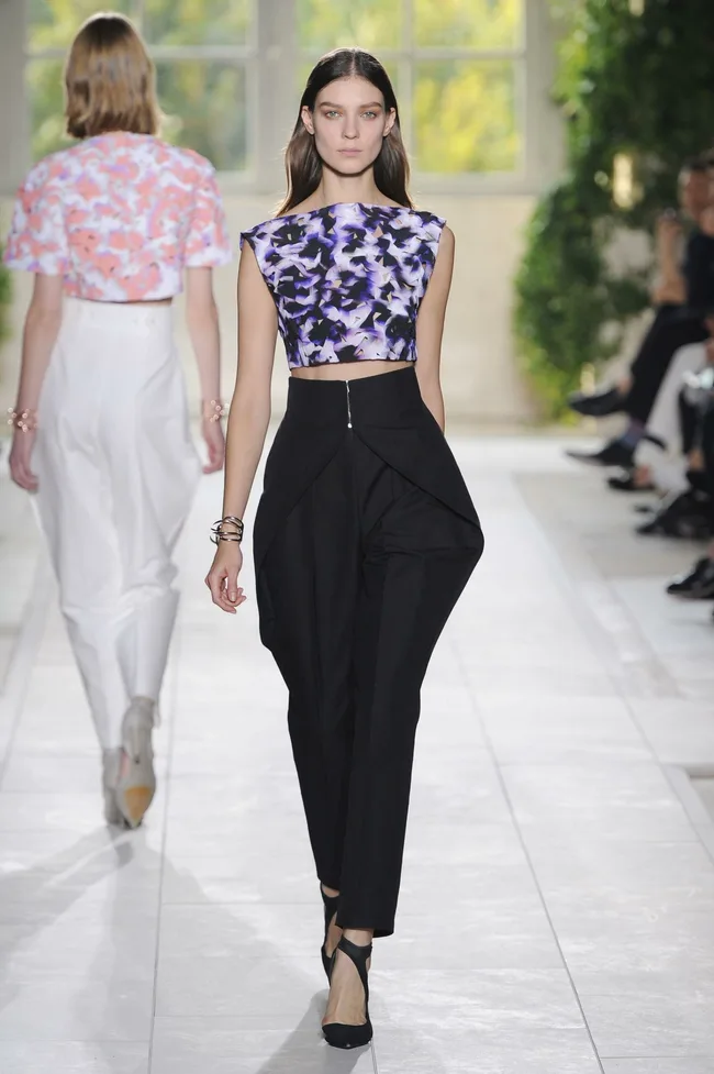 Fashion model walking the runway in a floral crop top and black pants during a daytime fashion show.