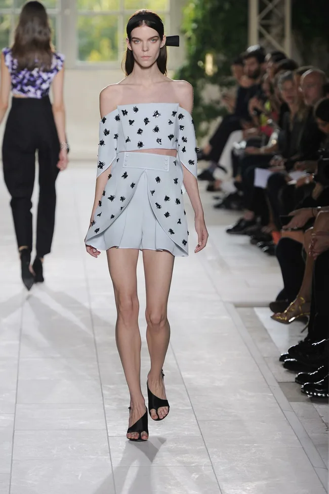 Model walking on a fashion runway wearing a light blue off-shoulder outfit with black floral patterns.