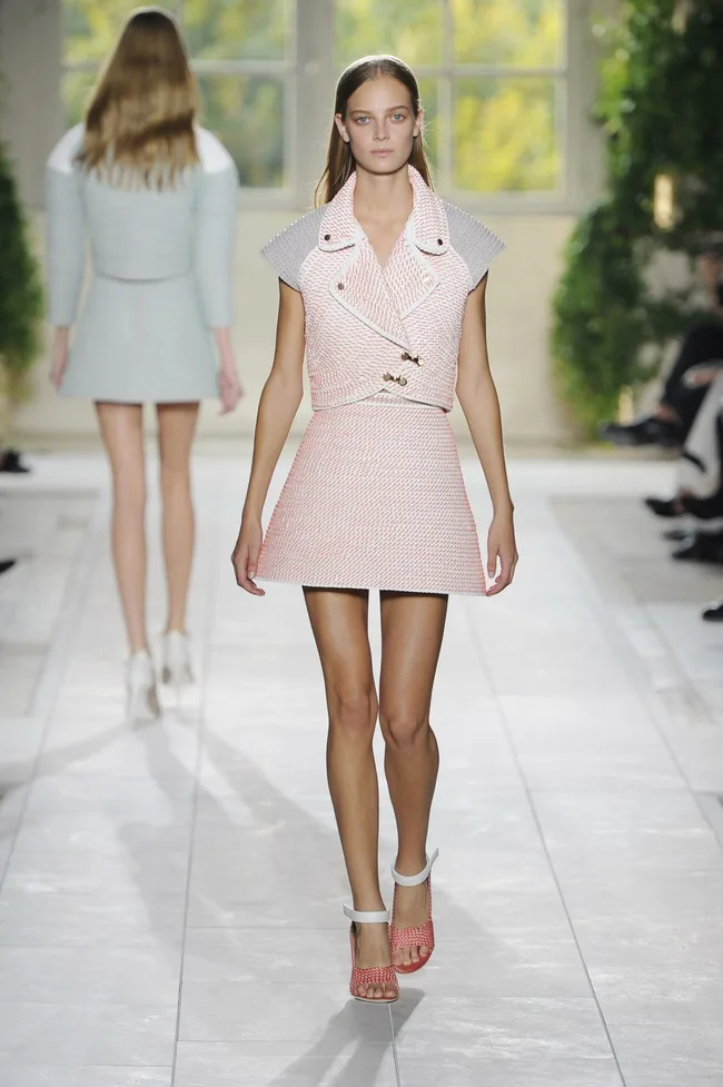 Model walking on runway in a structured pink and gray outfit with ankle strap sandals, runway environment in background.