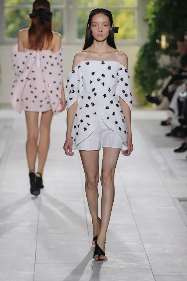 Model walks runway in a white off-shoulder dress with black floral patterns at a fashion show.