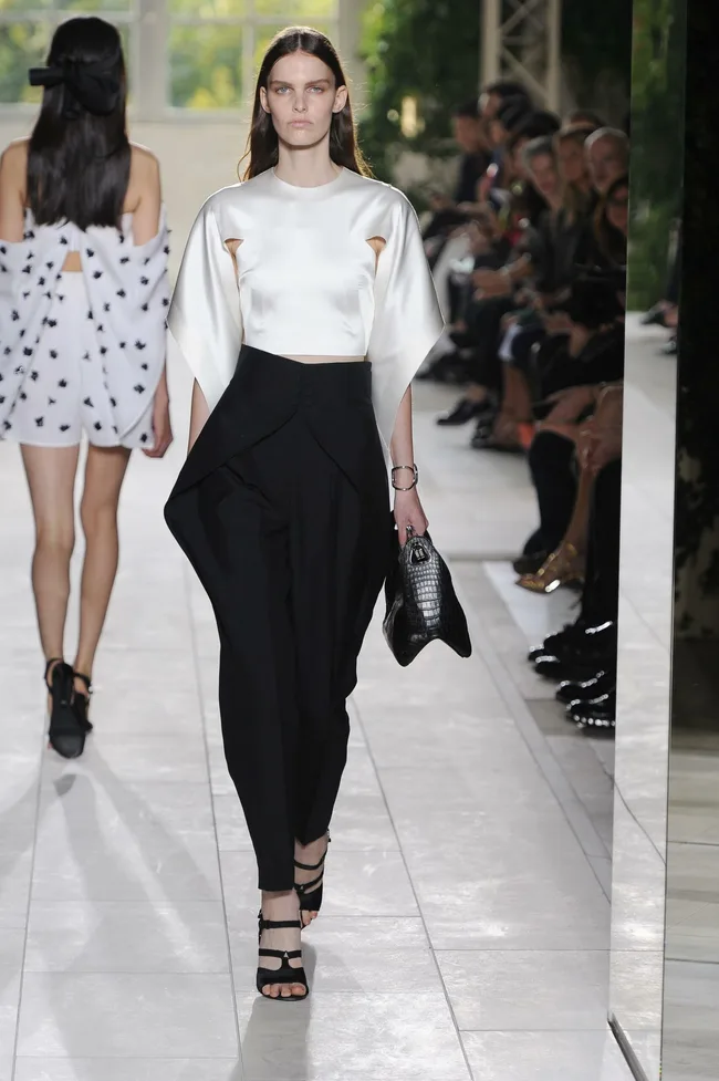 Model on runway in white blouse and black pants, holding a handbag. Fashion show audience in background.