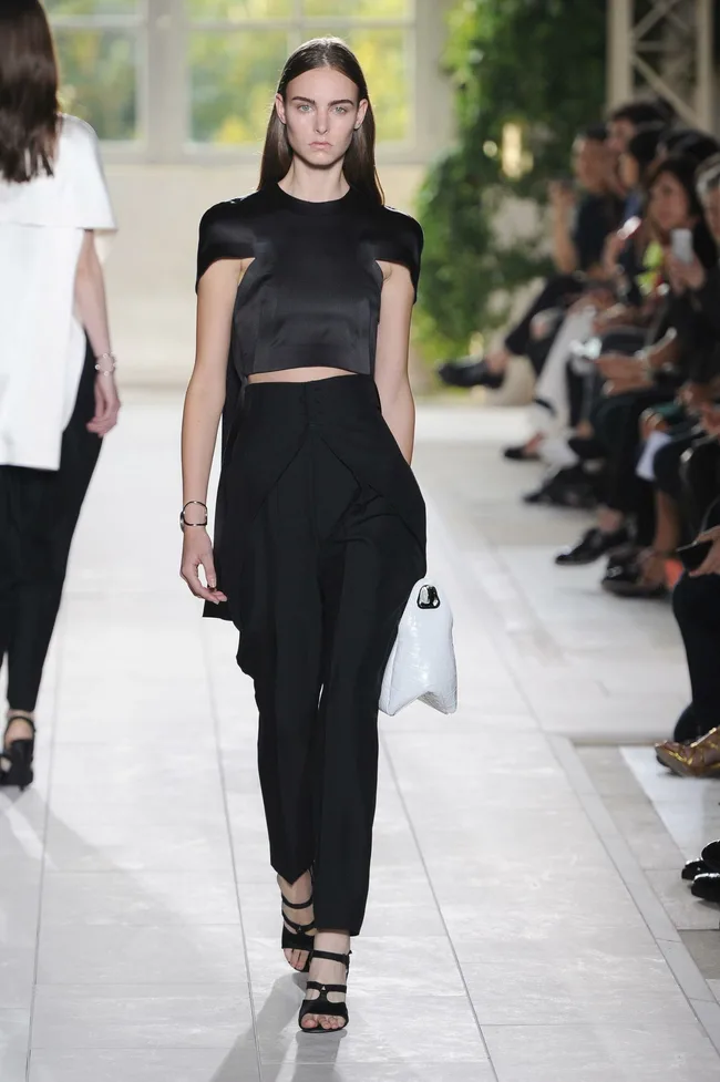 Model in a chic black outfit walks the runway at a fashion show, holding a white textured handbag.
