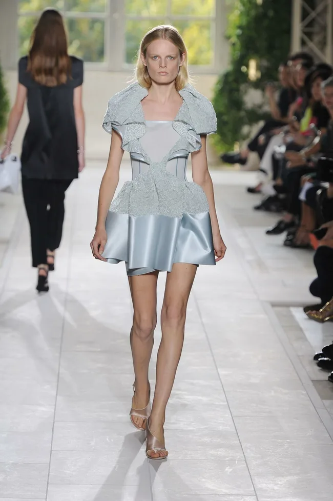 Model walking runway in light blue structured dress with ruffled details, surrounded by seated audience, blurred greenery.