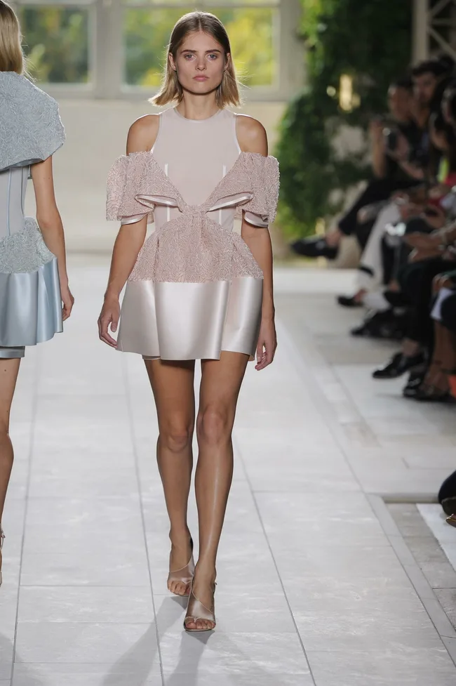 Model walks runway in a pink, structured dress at a fashion show, with spectators in the background.