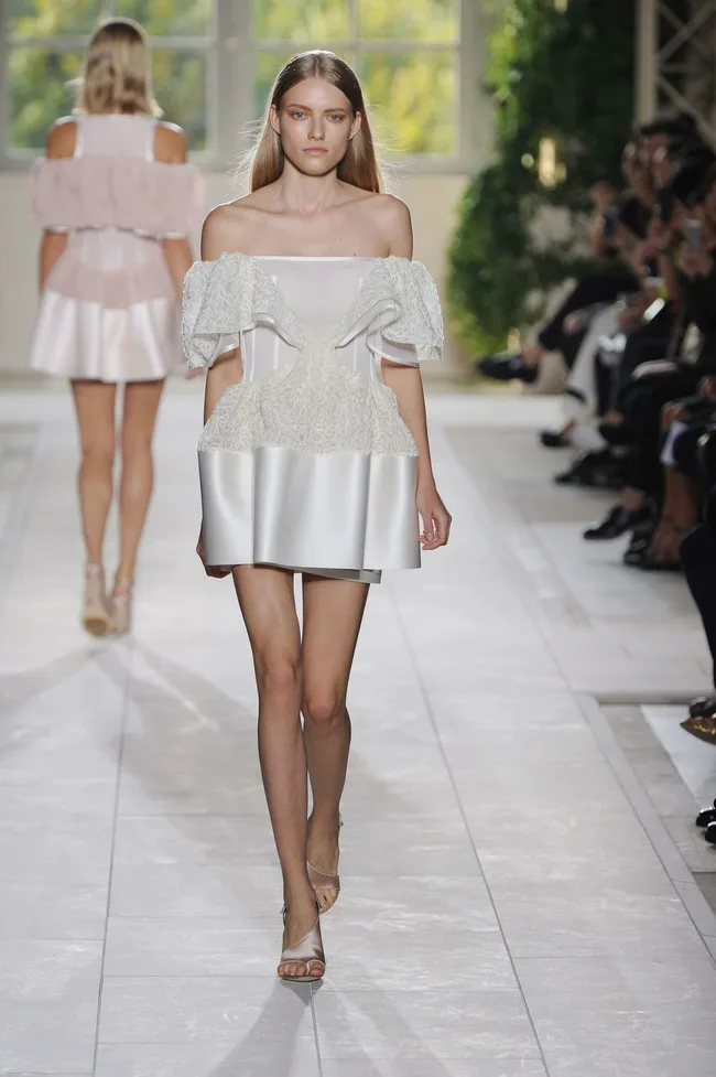 Model walking runway in a white off-shoulder, textured mini dress with an audience in the background.