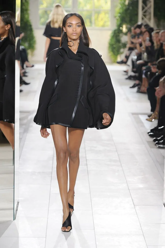 Model walking down a runway in a black, draped coat, with spectators in the background.