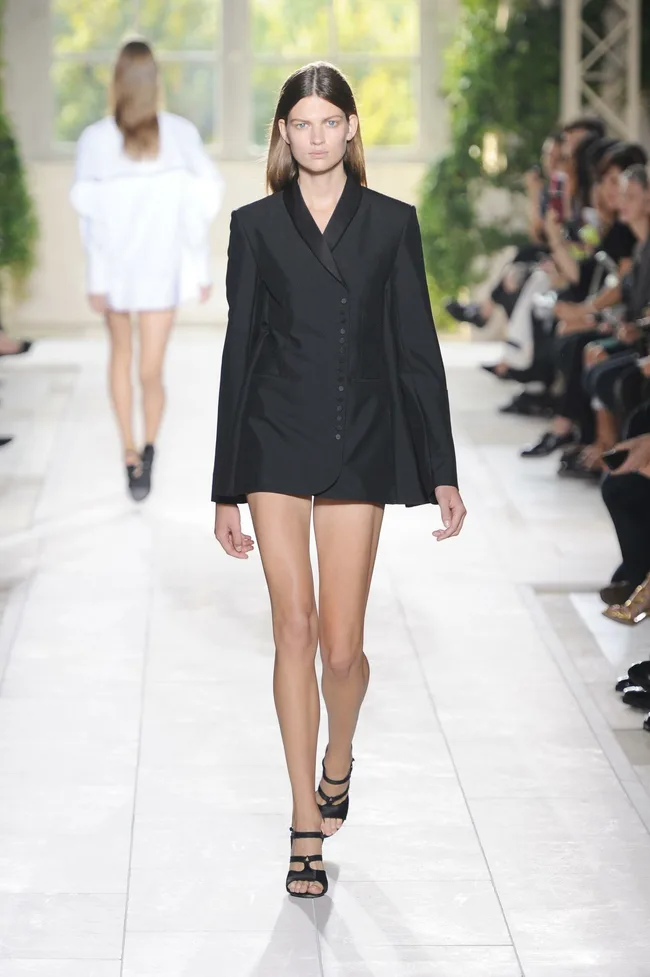 Model in black blazer dress walking down a runway with audience watching.