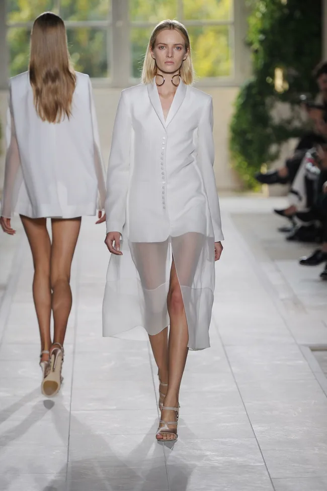 Model in white sheer outfit walking on a fashion runway.