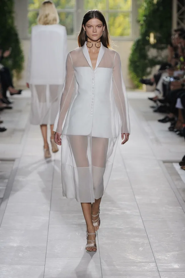 Model in a sheer white ensemble walking on a fashion runway, with onlookers in the background.