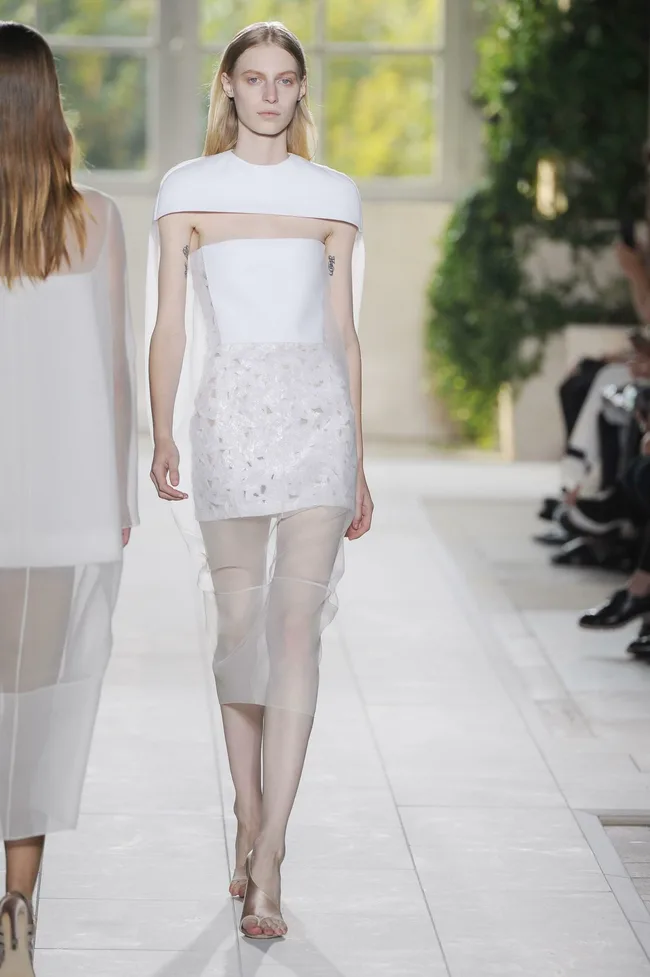 Model walks runway in a white outfit with sheer skirt at a fashion show, surrounded by greenery and audience.