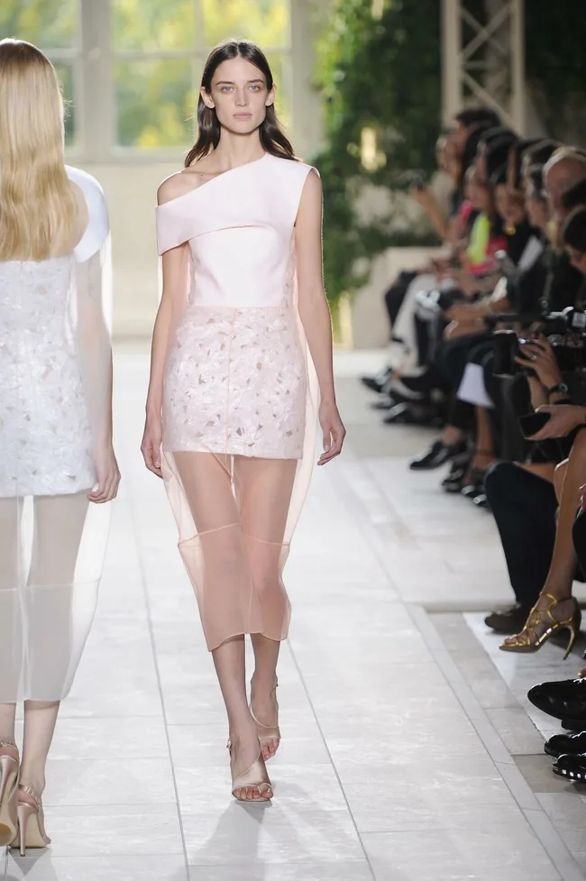 Model in a light pink asymmetrical dress walking the runway, surrounded by an audience in an indoor setting.