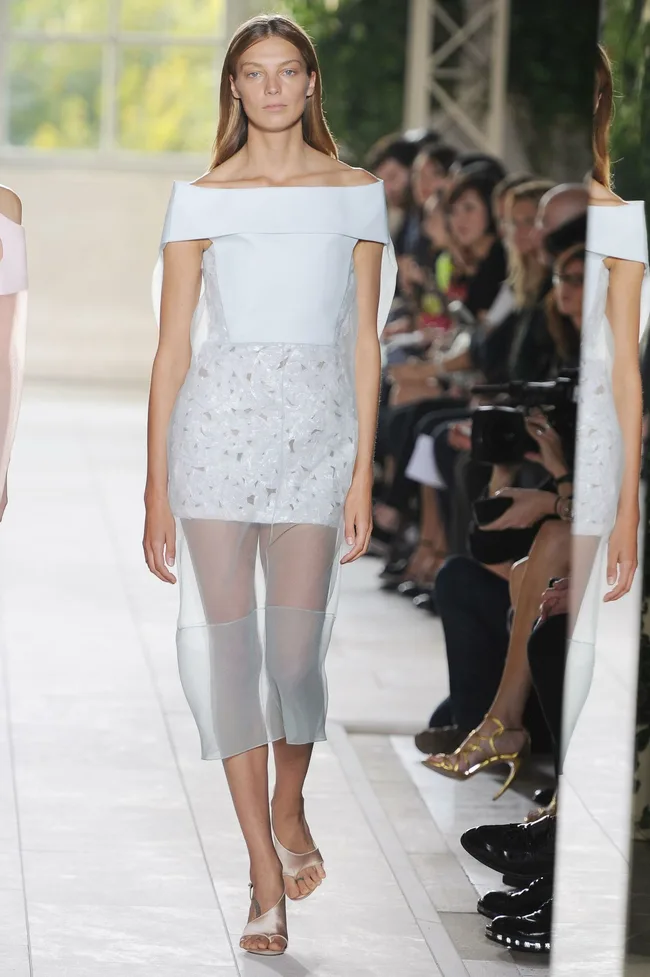 Model in a sheer white off-shoulder dress walks the runway, with an audience visible in the background.