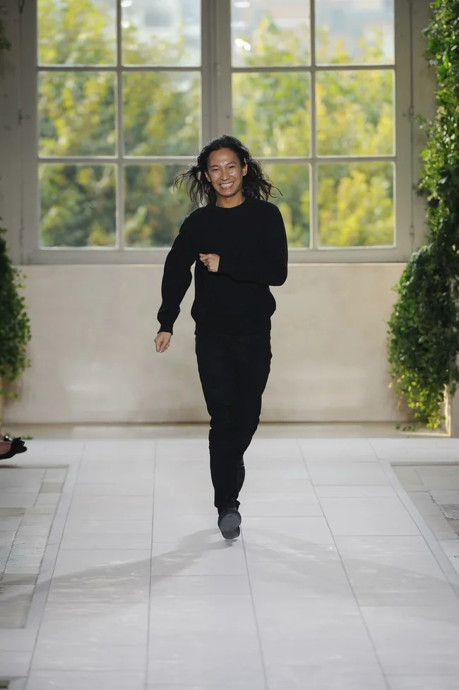Man in black outfit walking on a bright runway with greenery and large window backdrop.