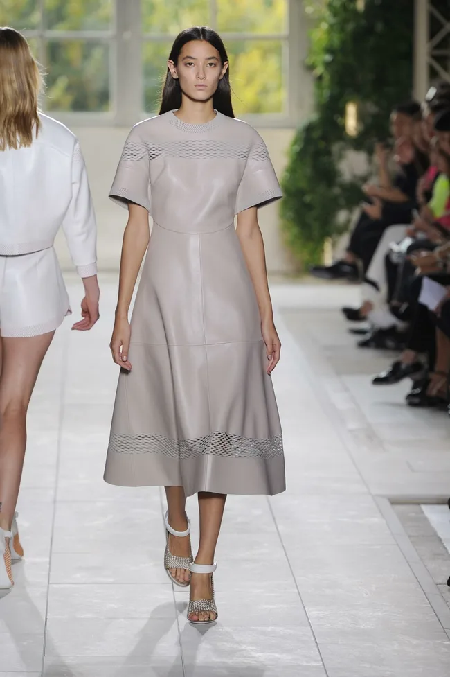 Model walks runway in a light gray, short-sleeved leather dress with netted details, at a fashion show.