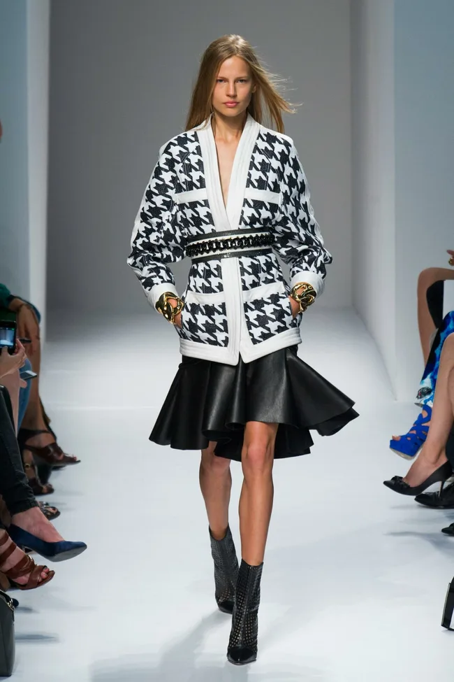 Model walks runway in black and white houndstooth jacket with flared black skirt and ankle boots.