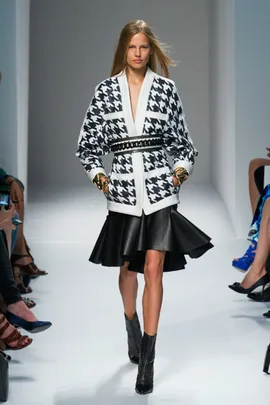 Model walks runway in black and white houndstooth jacket with flared black skirt and ankle boots.