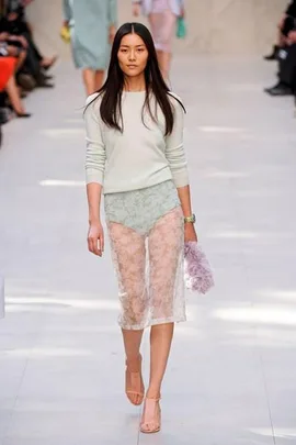 Model walking on a runway wearing a sheer lace skirt and pastel top, holding a fluffy handbag, with audience in the background.