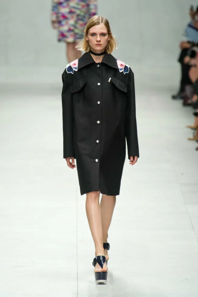 Model in black button-up coat with floral shoulder details, walking on a runway at Carven SS14 fashion show.