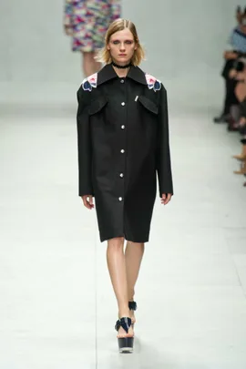 Model in black button-up coat with floral shoulder details, walking on a runway at Carven SS14 fashion show.