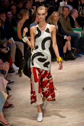 Model walking runway in a white dress with black and red abstract patterns, in front of an audience during a fashion show.