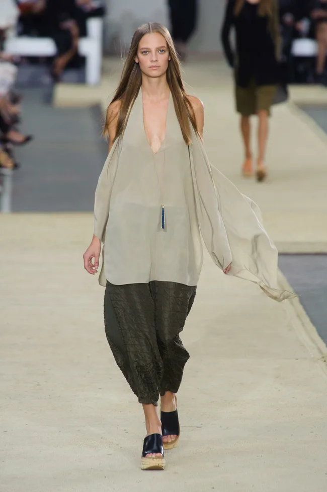 Model walks runway in sheer beige halter top and dark loose pants during fashion show.